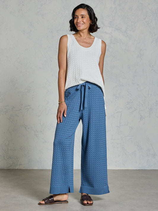 Woman wearing a white sleeveless top and blue patterned pants against a gray background