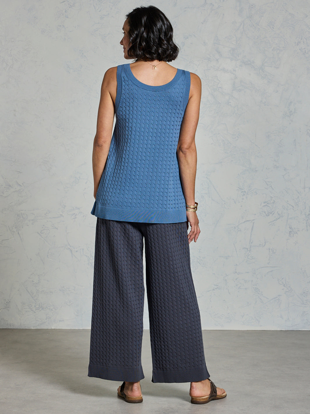 Woman wearing a blue sleeveless top and dark pants against a gray background