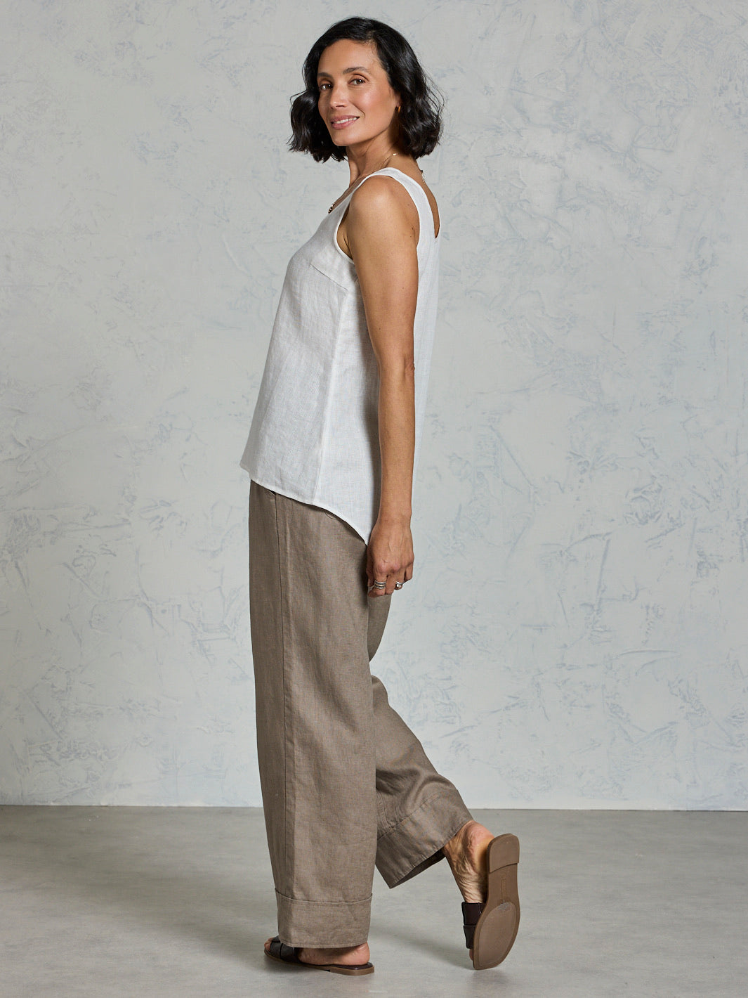 Woman wearing a white sleeveless top and brown pants against a gray background