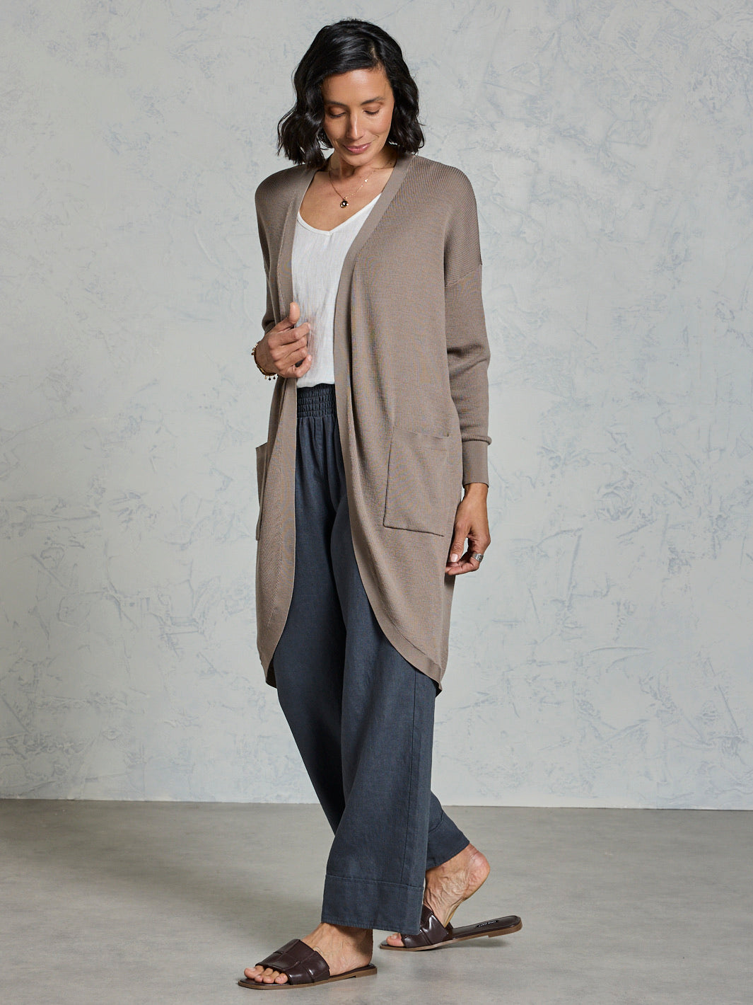 Woman wearing a long beige cardigan over a white top and dark pants against a plain background