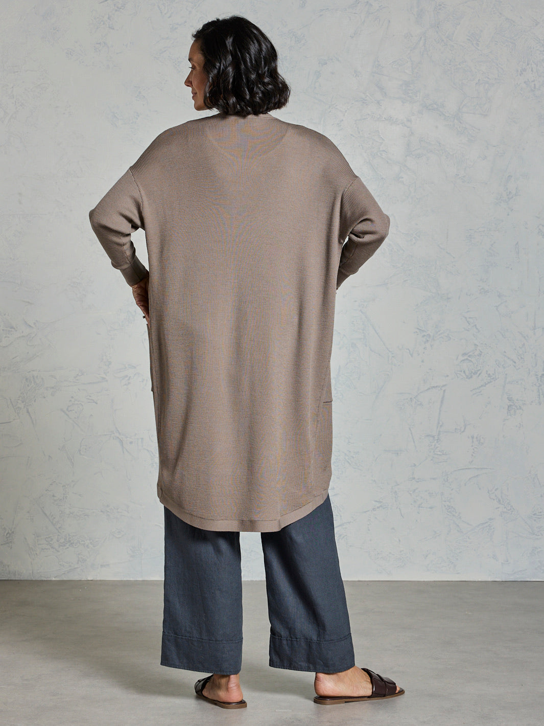 Person wearing a long beige cardigan and dark pants against a plain background