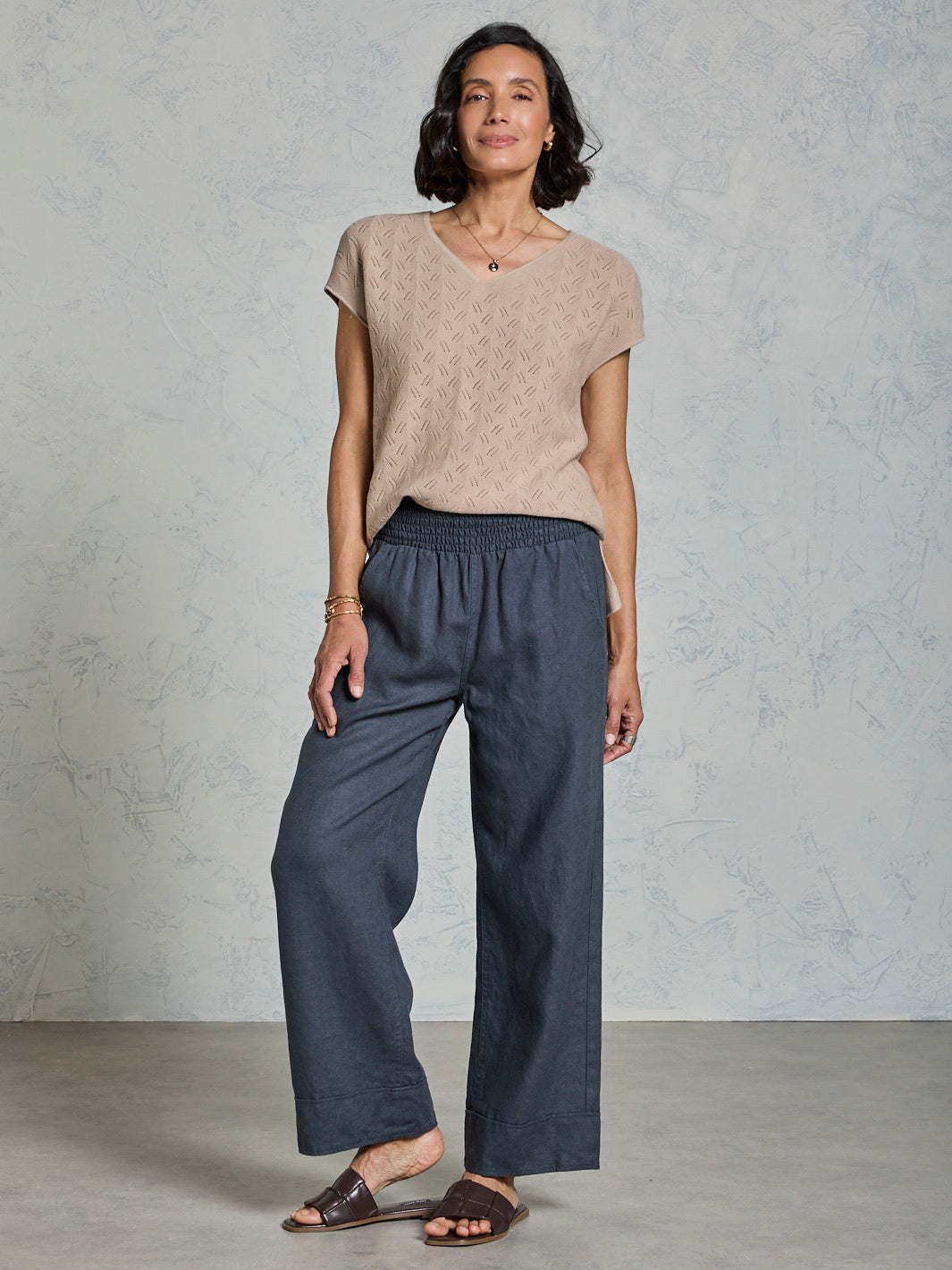 Woman wearing a beige top and dark gray pants against a plain background