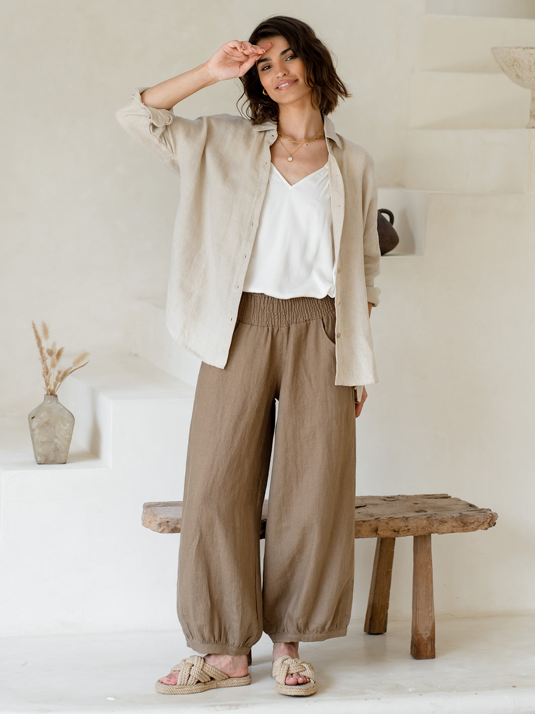 A female model standing in a room with a neutral backdrop, wearing beige French linen pants with a relaxed fit and tapered hem, paired with a white top and beige flat shoes.