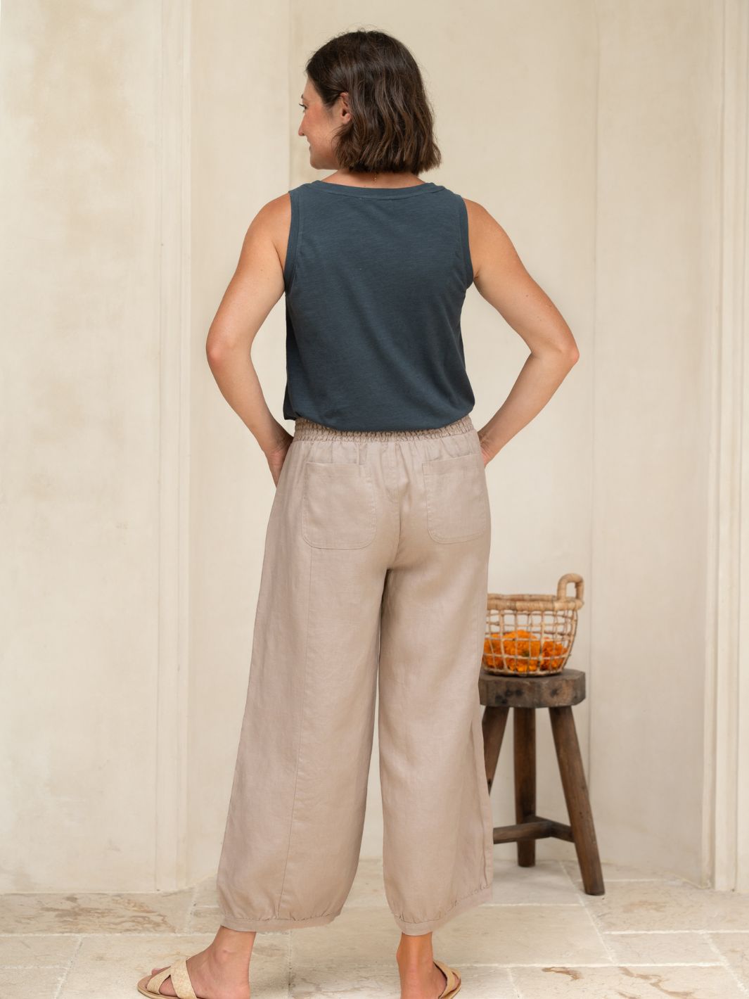 Woman wearing a sleeveless top and wide-leg pants standing in a room with a wooden stool and basket.