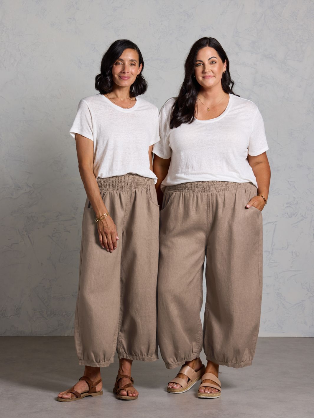 Two women wearing white tops and brown wide-leg pants standing against a plain background