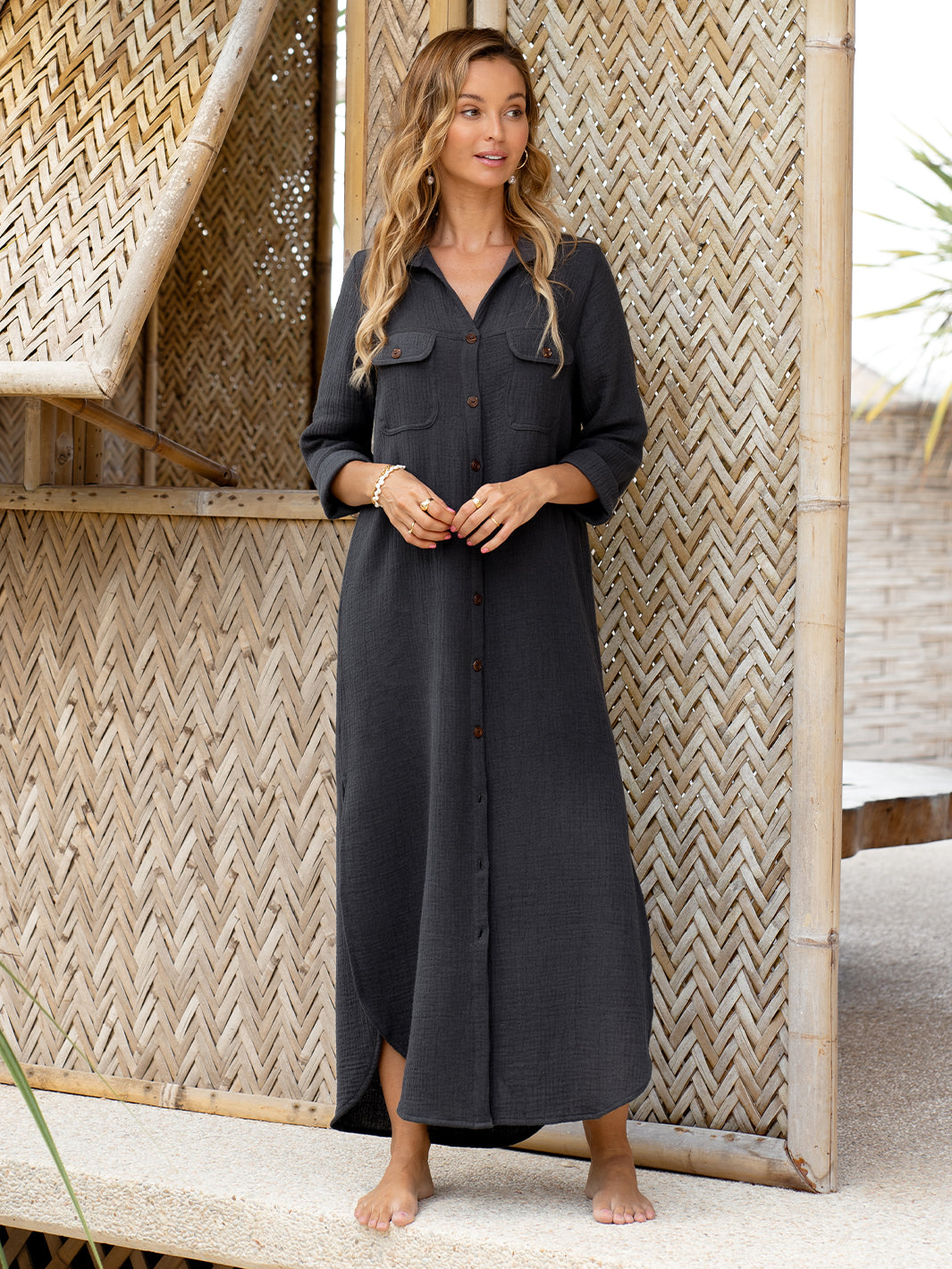 A woman standing in front of a bamboo wall, wearing a long charcoal-colored linen blend shirt dress with a curved hemline, patch pockets, and natural coconut buttons.