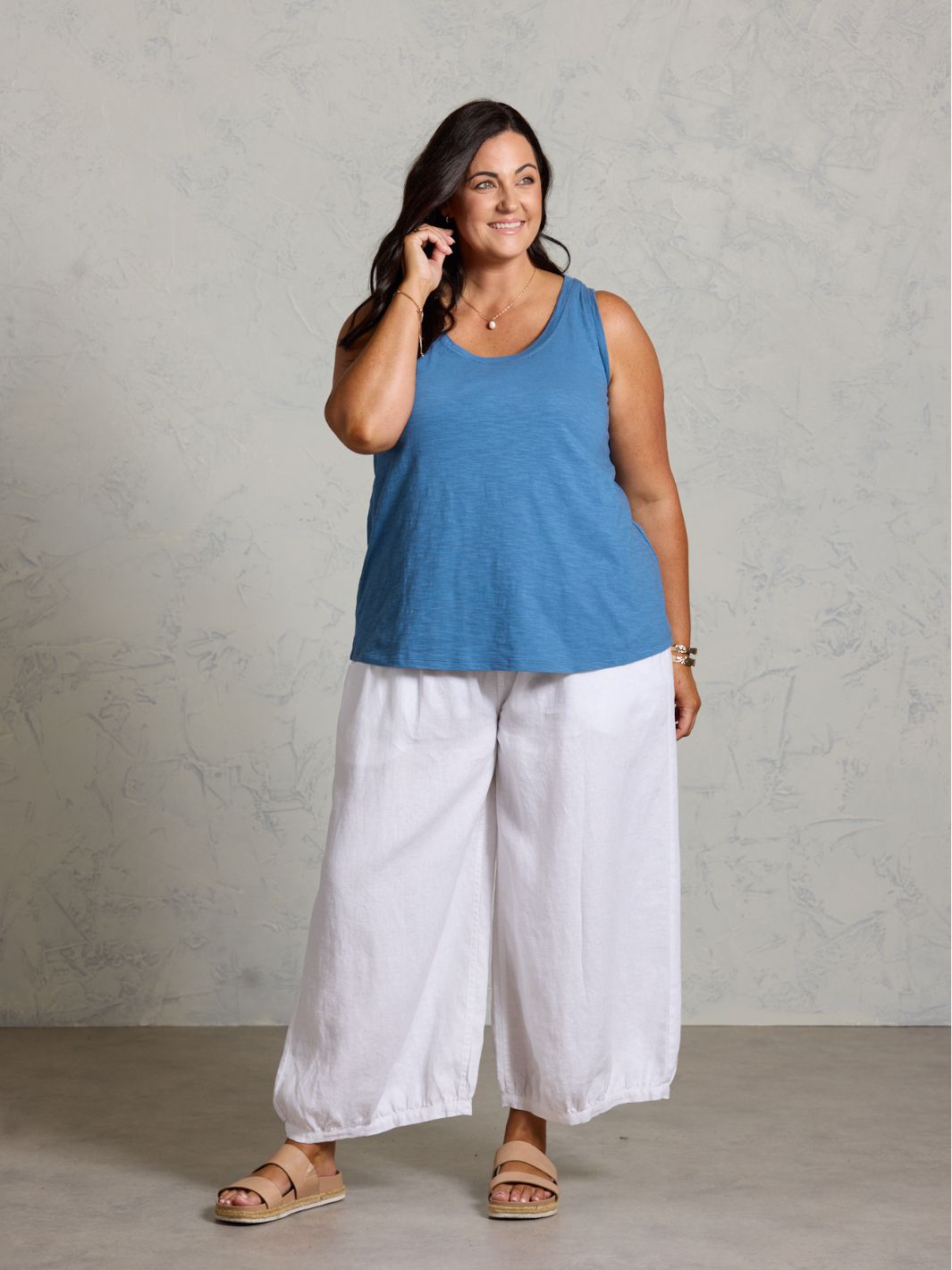 Woman wearing a blue sleeveless top and white pants against a gray background