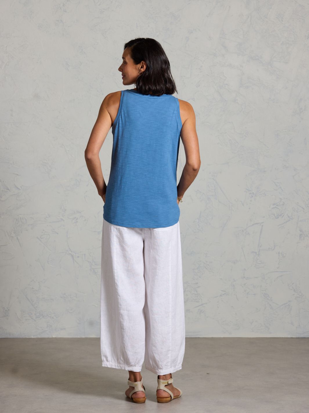 Woman wearing a blue tank top and white pants against a gray background