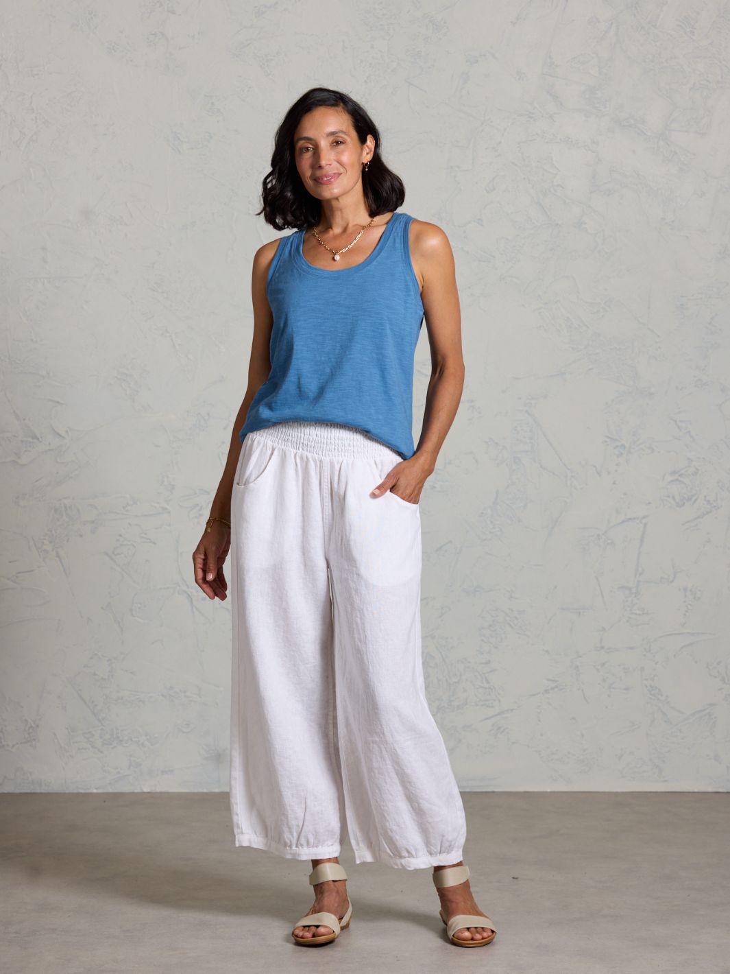 Woman wearing a blue sleeveless top and white pants against a plain background