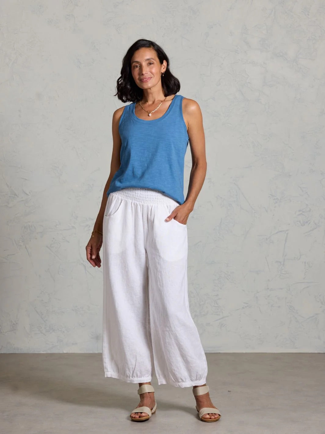 Woman wearing a blue sleeveless top and white pants against a gray background