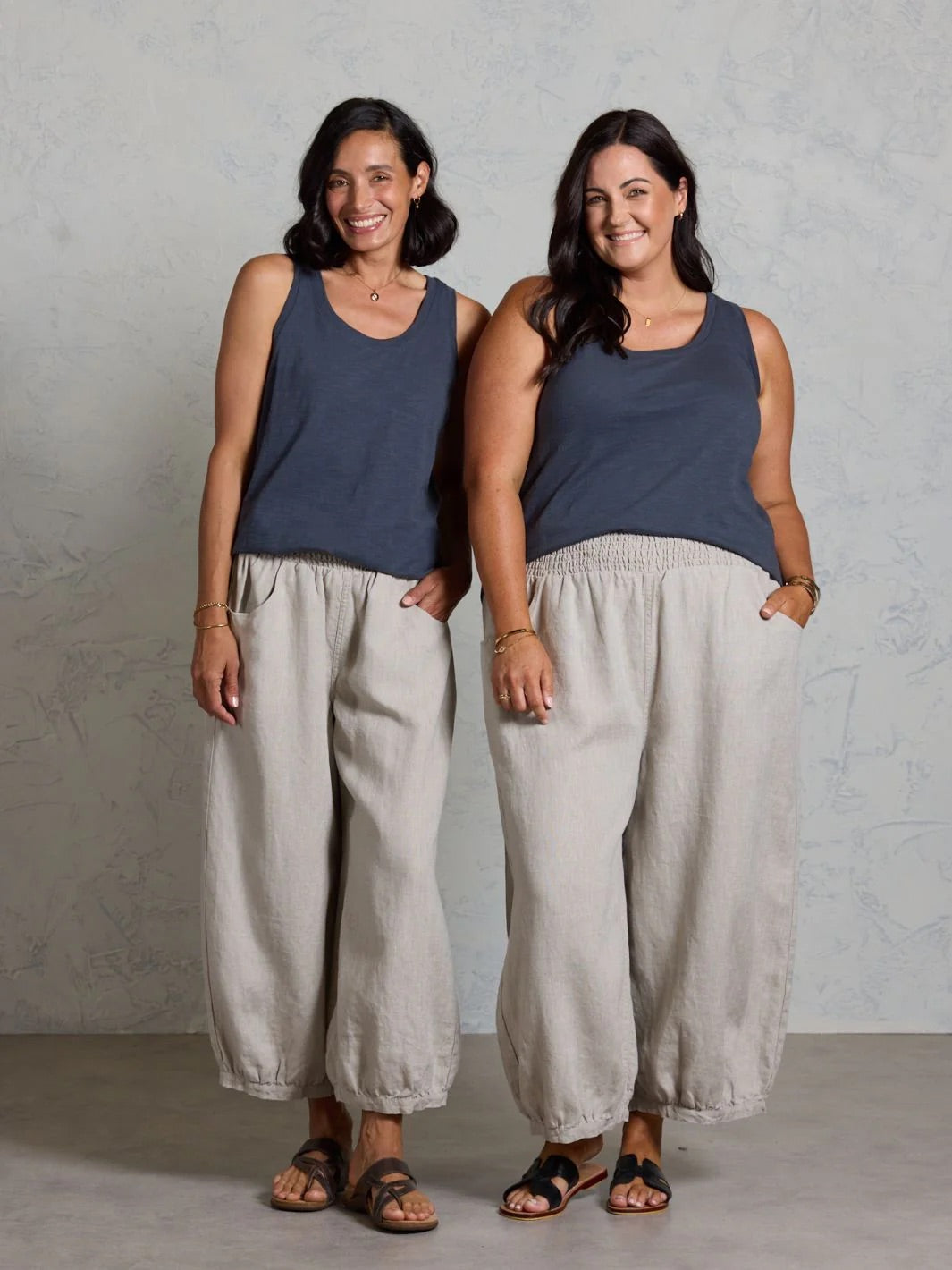 Two women wearing matching black tank tops and beige pants against a plain background