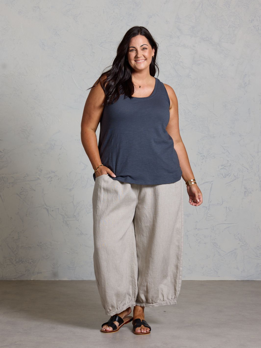 Woman wearing a dark tank top and light gray pants against a plain background