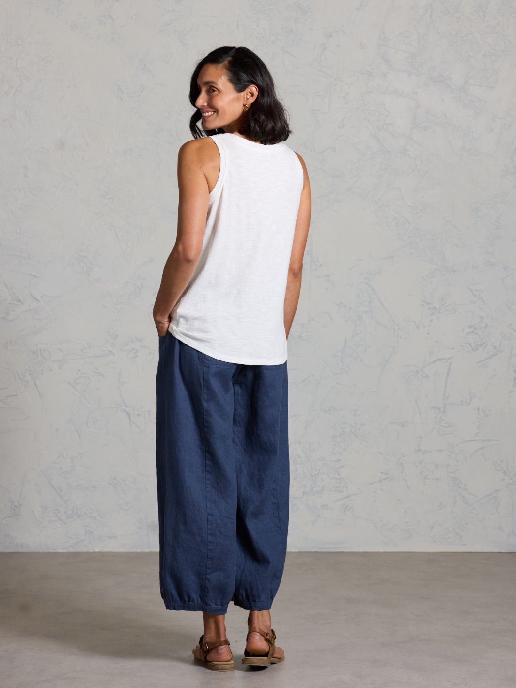 Woman wearing a white sleeveless top and dark pants against a plain background