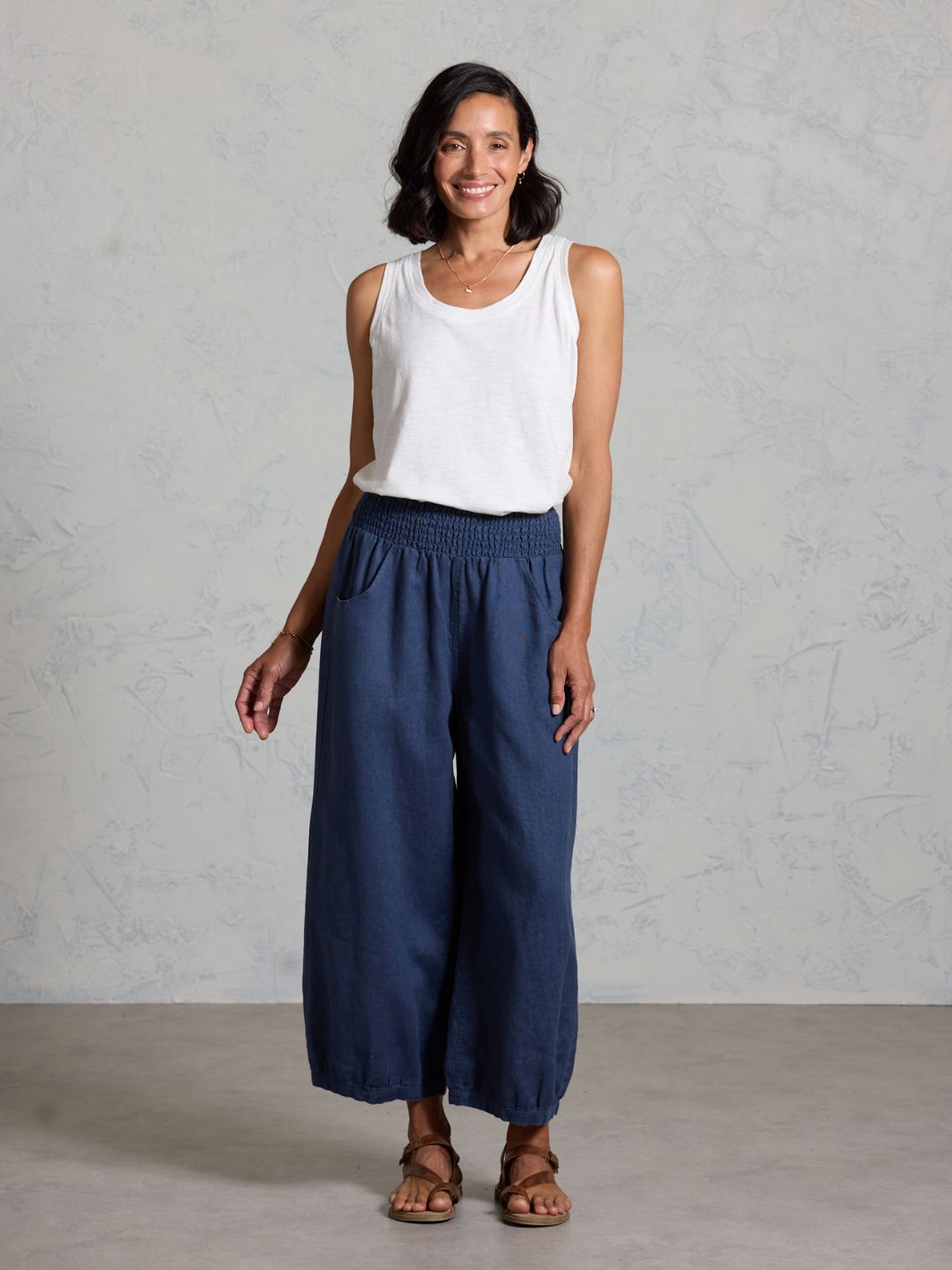 Woman wearing a white sleeveless top and dark wide-leg pants against a gray background