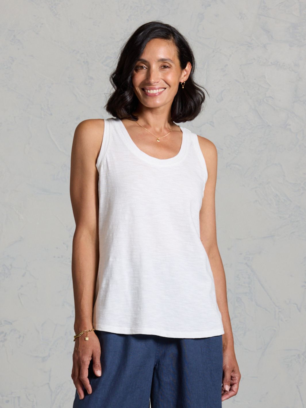 Woman wearing a white tank top and dark pants against a gray background