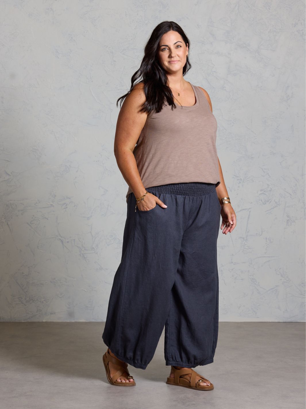 Woman wearing a brown tank top and dark wide-leg pants against a gray background