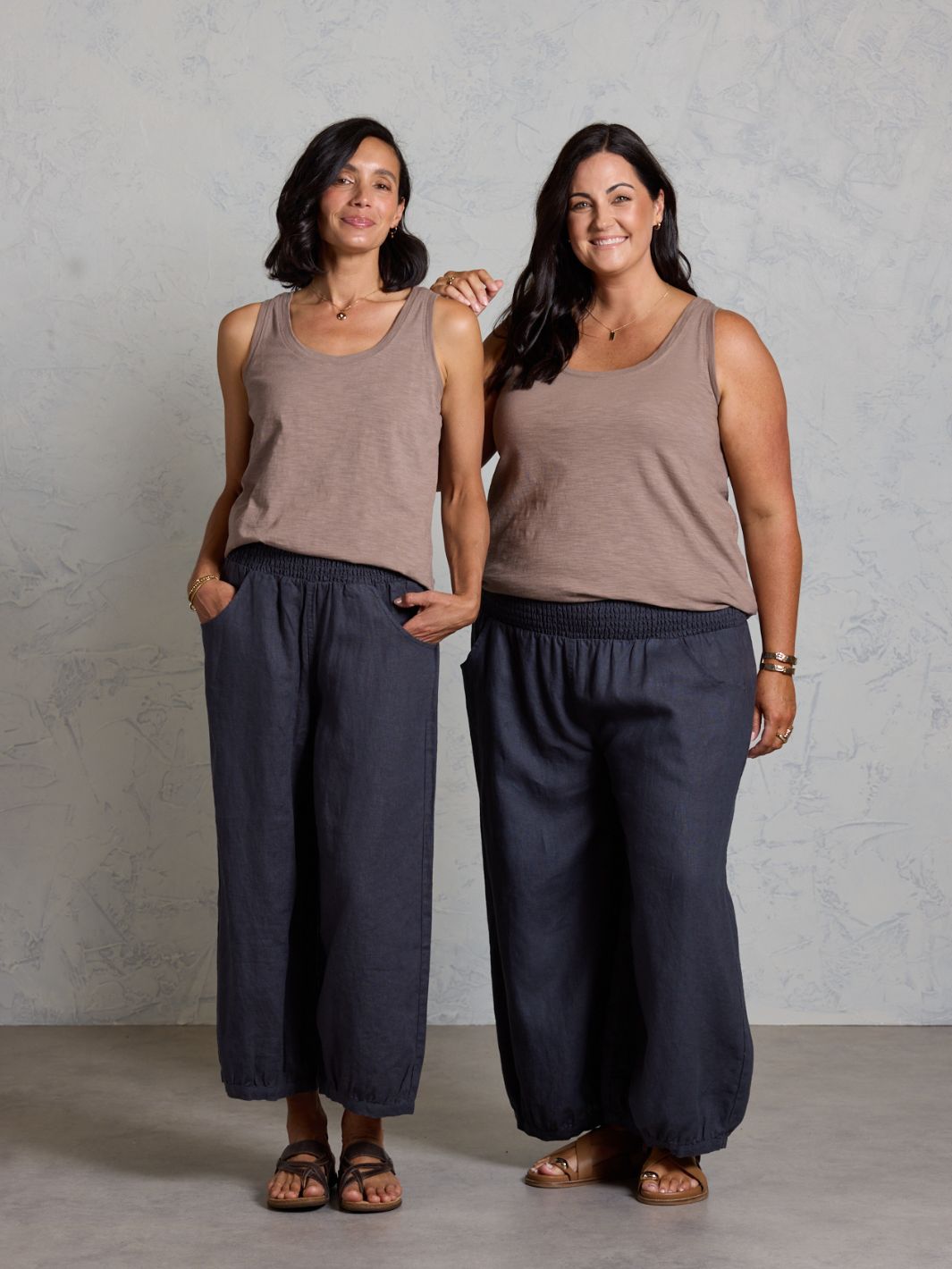 Two women wearing matching brown tank tops and dark pants against a gray background