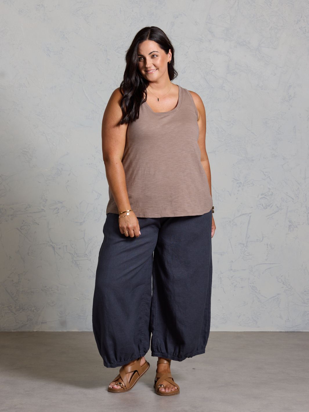 Woman wearing a brown tank top and dark wide-leg pants against a gray background