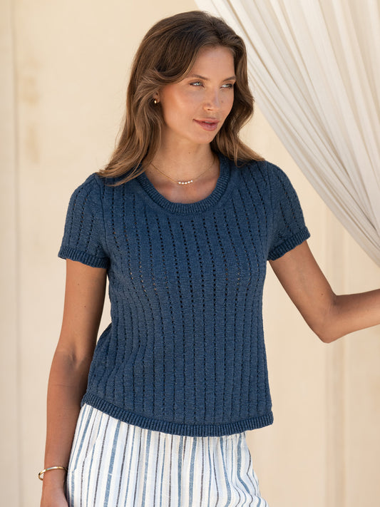 Model wearing the Agni Cotton Linen Knit Top in navy, styled with striped linen pants in a sunlit doorway.