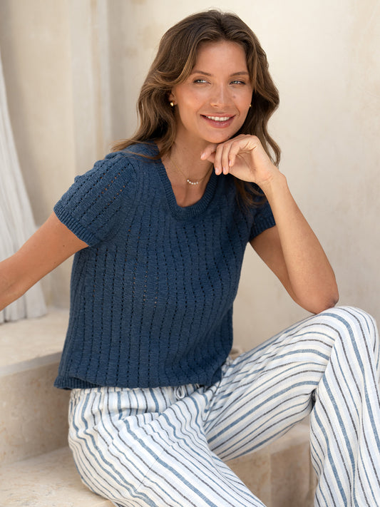 Model seated in the Agni Cotton Linen Knit Top in navy with striped linen pants, smiling in soft natural light.