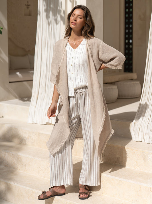 Model wearing the Laguna French Linen Kimono in natural with a white top and striped linen pants, on stone steps in dappled sunlight.