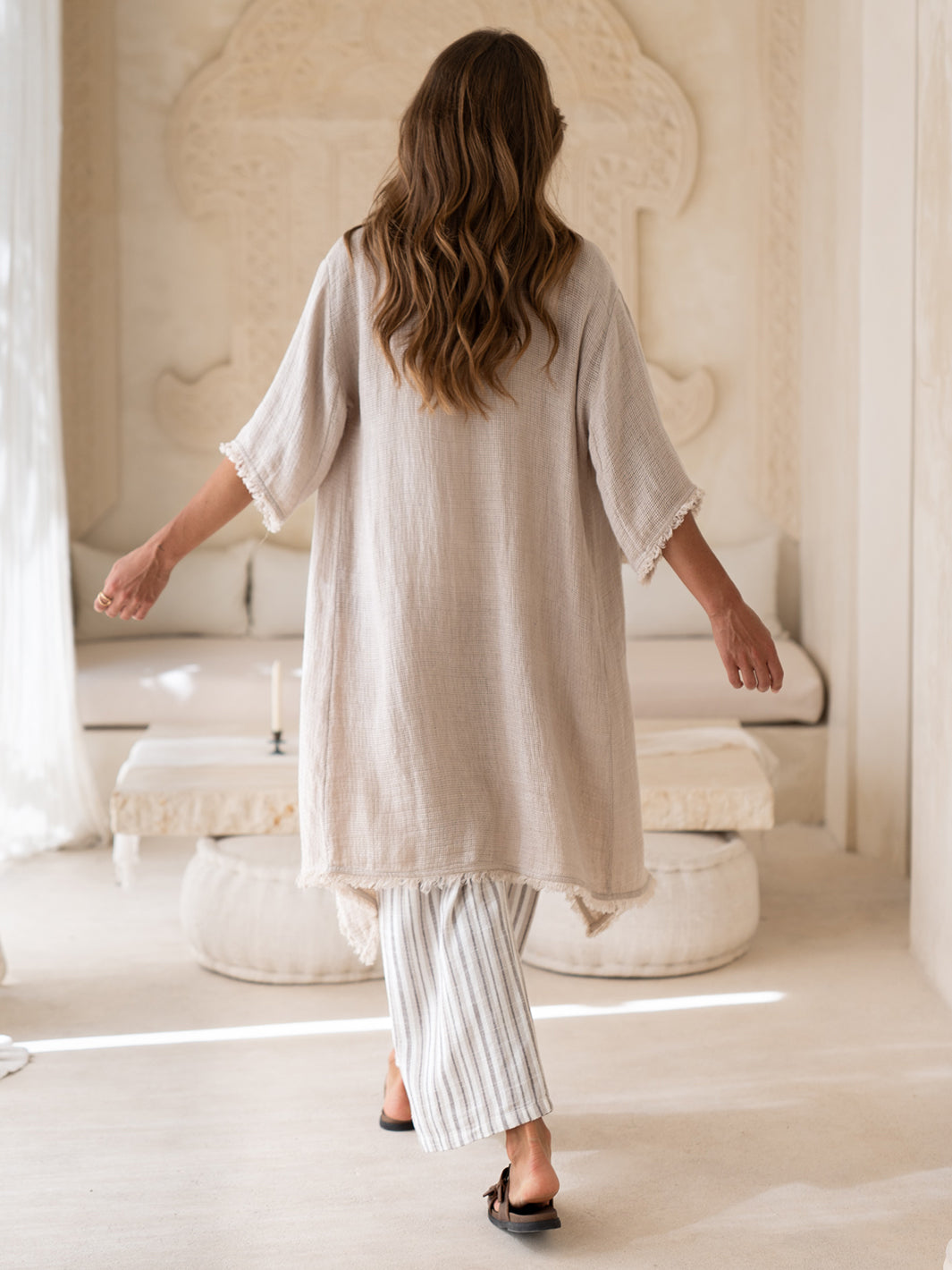 Back view of the Laguna French Linen Kimono in natural in motion, with striped linen pants in a sunlit interior.