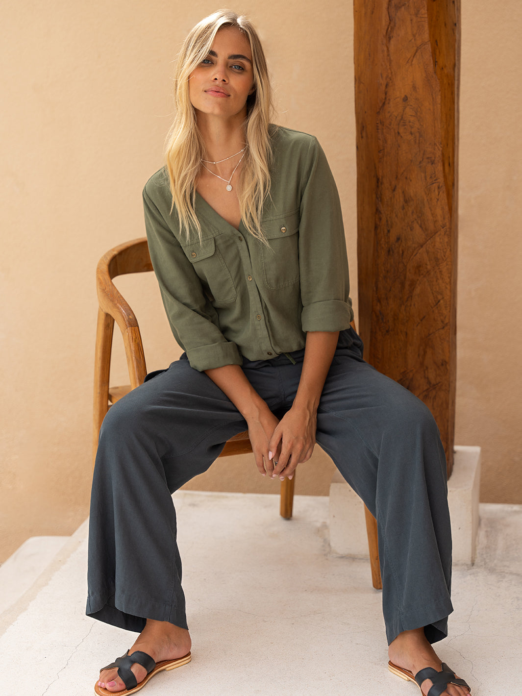 A woman sitting on a chair wearing an olive green Yves Cargo Top with cargo patch pockets, full-length sleeves, and a V-neckline.